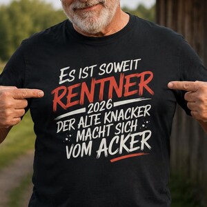 Könnte beinhalten: Ein schwarzes Kurzarm-T-Shirt mit weißem und rotem Text: Es ist soweit RENTNER 2026 DER ALTE KNACKER MACHT SICH VOM ACKER. Das grafische Design ist mittig auf der Brust des T-Shirts platziert, das mit einer blauen Jeanshose getragen wird.