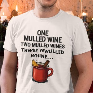 Könnte beinhalten: Weißes T-Shirt mit dem Text "ONE MULLED WINE TWO MULLED WINES THWEE MWULLED WHINE..." und einer Grafik einer roten Tasse mit Glühwein. Die Person steht vor einem weihnachtlich geschmückten Kamin.
