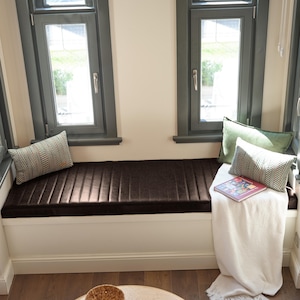May include: A window seat with a dark brown cushion and two decorative pillows. One pillow has a chevron pattern, and the other is solid green. A book and a white blanket are also on the seat.