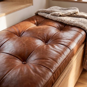 May include: A rectangular bench featuring a tufted brown leather cushion with visible button detailing. A thick, textured beige knit throw blanket is draped over the right side of the leather seat, which rests upon a light-toned wooden base structure.