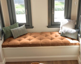 May include: A long, rectangular, tufted bench cushion in a solid tan color, positioned on a white window seat. The cushion is accented with two solid green pillows and two patterned pillows featuring a white and dark green chevron design.