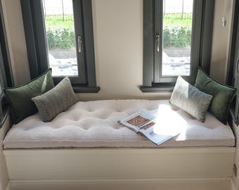 Könnte beinhalten: Eine weiße Fensterbank mit einem getufteten cremefarbenen Polster, zwei einfarbigen dunkelgrünen rechteckigen Kissen und zwei gemusterten Fischgrätkissen. Ein aufgeschlagenes Buch liegt auf dem Polster zwischen den dunkelgrauen Fensterrahmen.