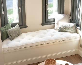 Puede incluir: Un cojín de banco largo y blanco con capitoné se asienta sobre un asiento de ventana integrado. El cojín está acentuado por una almohada cuadrada de color verde oliva, una rectangular con patrón de espiga verde y blanco y una blanca.