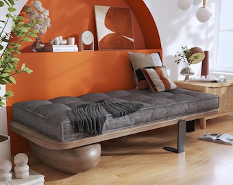 May include: A low-profile daybed featuring a dark gray tufted cushion on a light wood frame with a rounded base and a black metal support leg. A dark gray throw blanket is draped over the cushion, accompanied by two square patterned throw pillows.