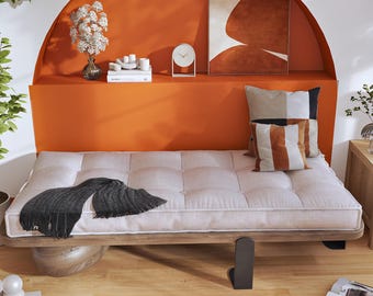 Puede incluir: Un diván de perfil bajo con un cojín beige capitoné sobre una estructura de madera, que incluye una manta gris oscuro con flecos, un cojín cuadrado de bloques de color y un cojín rectangular a rayas en tonos marrón, blanco y negro.