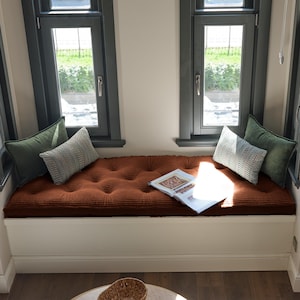 May include: A white window seat bench featuring a tufted rust-colored cushion. The bench is adorned with two solid dark green rectangular pillows and two patterned gray and white chevron pillows. An open book rests on the cushion surface.