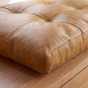 May include: A square tan leather floor cushion featuring a tufted design with visible stitching and button accents. The surface of the cushion displays a natural grain texture and a soft, matte finish, showcasing the detailed craftsmanship.
