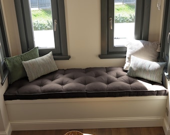 May include: A long, rectangular dark gray tufted bench cushion sits on a white window seat. The cushion is accented with two green velvet pillows, two patterned gray and white rectangular pillows, and one solid light gray square pillow.
