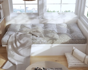May include: A rectangular window seat cushion featuring a tufted design with a muted green and white floral pattern. A grey and white herringbone throw blanket is draped over the cushion. Two rectangular white pillows with thin horizontal stripes.