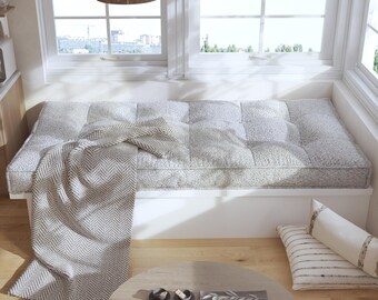 Puede incluir: Un cojín rectangular para ventana tapizado en tela gris claro texturizada con diseño capitoné. Una manta con patrón de espiga en beige y blanco está colocada sobre el cojín. Dos cojines rectangulares con rayas horizontales se encuentran al lado.