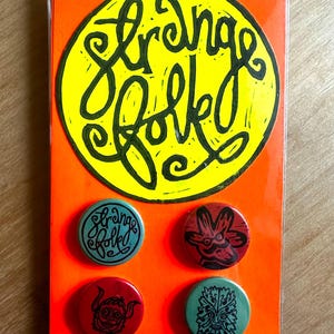 May include: An orange card featuring a yellow circle with the words "Strange Folk" in black script. Four illustrated round badges are displayed on the card, with the words "Strange Folk".