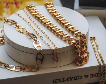May include: A collection of gold chains, including a thick curb chain, a delicate chain, and a chain with a unique link design. The chains are displayed on a white surface with a book titled "Made & Crafted".