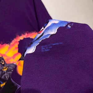 Puede incluir: Camiseta morada con un gr&aacute;fico de Batman. El dise&ntilde;o muestra a Batman con un traje negro y el s&iacute;mbolo del murci&eacute;lago amarillo, sobre un fondo de fuego. La camiseta incluye el texto "&copy;1991 DC Comics Inc."
