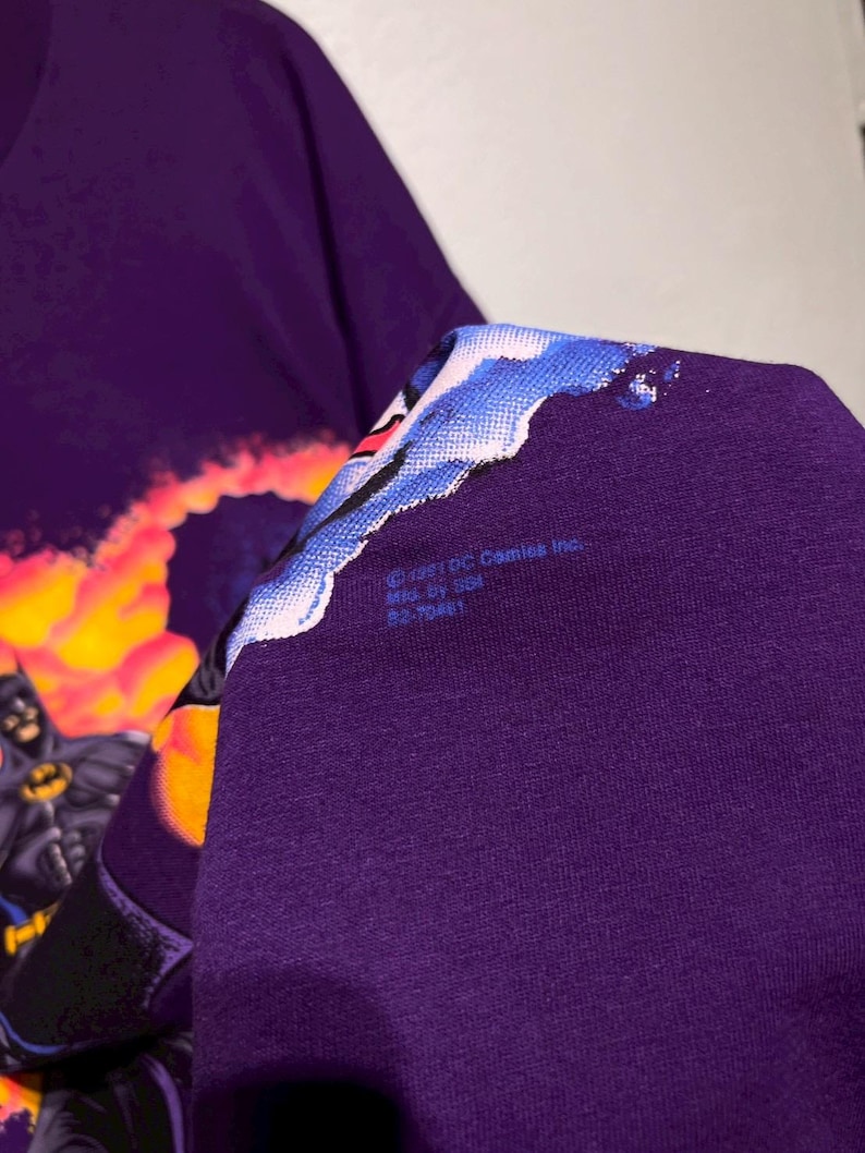 Puede incluir: Camiseta morada con un gr&aacute;fico de Batman. El dise&ntilde;o muestra a Batman con un traje negro y el s&iacute;mbolo del murci&eacute;lago amarillo, sobre un fondo de fuego. La camiseta incluye el texto "&copy;1991 DC Comics Inc."