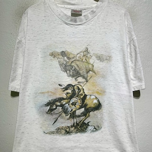 Peut inclure: T-shirt blanc moucheté avec une illustration de chasse au bison par des Amérindiens à cheval. L'œuvre, aux tons sépia, représente des personnages, des chevaux et des bisons, capturant une scène historique.