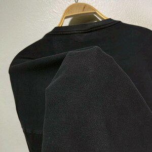 May include: A black crew neck sweatshirt is displayed on a wooden hanger. The fabric is folded, revealing the texture of the material. The sweatshirt is hanging against a light-colored wall.