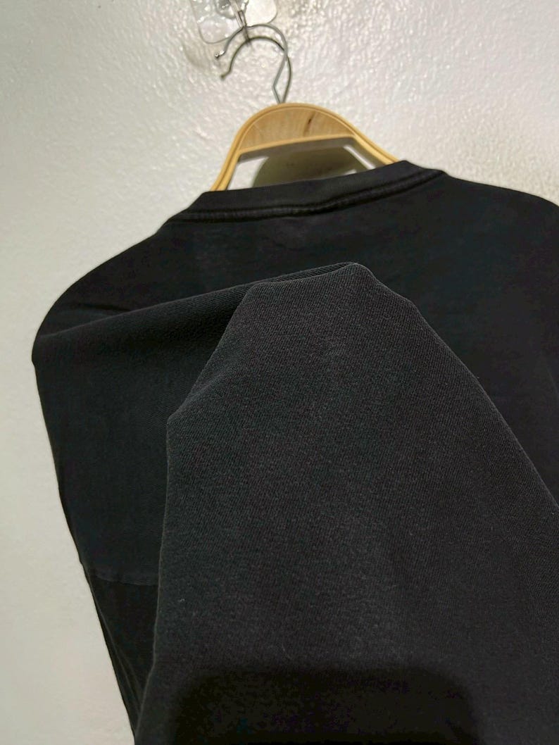 May include: A black crew neck sweatshirt is displayed on a wooden hanger. The fabric is folded, revealing the texture of the material. The sweatshirt is hanging against a light-colored wall.