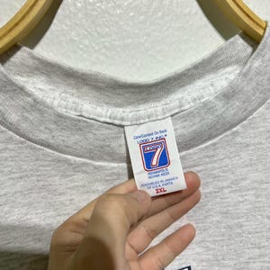 May include: A light gray t-shirt with a crew neck. The tag reads "LOGO 7, INC" and "INDIANAPOLIS INDIANA 46226". The shirt is a 2XL size. The shirt is hanging on a wooden hanger.