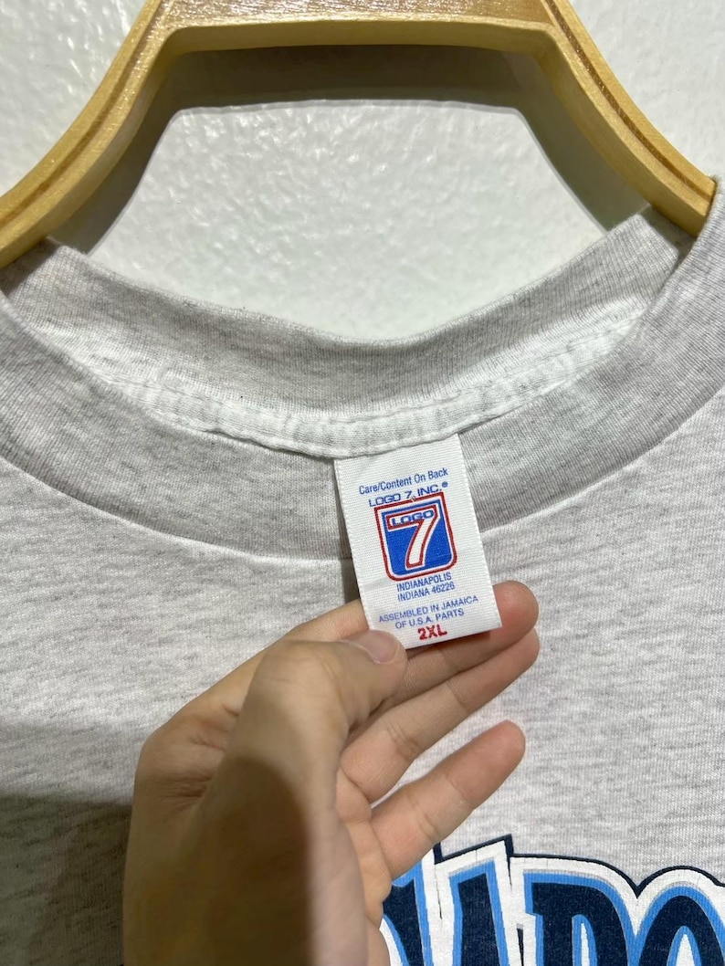 May include: A light gray t-shirt with a crew neck. The tag reads "LOGO 7, INC" and "INDIANAPOLIS INDIANA 46226". The shirt is a 2XL size. The shirt is hanging on a wooden hanger.