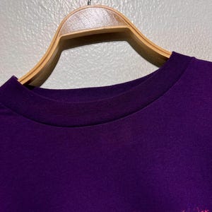 Puede incluir: Primer plano de una camiseta morada con cuello redondo, colgada de una percha de madera. La parte inferior de la camiseta presenta un gr&aacute;fico colorido con tonos de rosa, naranja y amarillo.