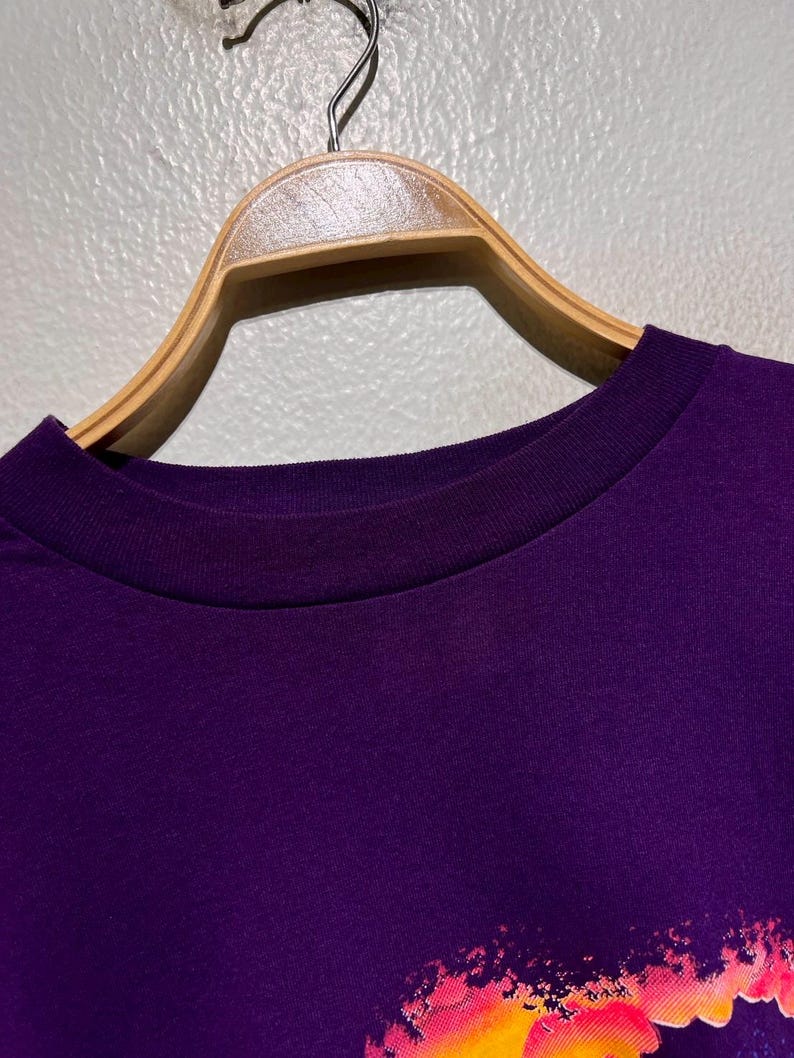 Puede incluir: Primer plano de una camiseta morada con cuello redondo, colgada de una percha de madera. La parte inferior de la camiseta presenta un gr&aacute;fico colorido con tonos de rosa, naranja y amarillo.