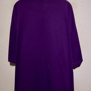 Puede incluir: Mono morado liso con cuello alto y mangas cortas, colgado de una percha de madera. La prenda se exhibe contra una pared neutra. El mono parece estar hecho de un material suave y c&oacute;modo.