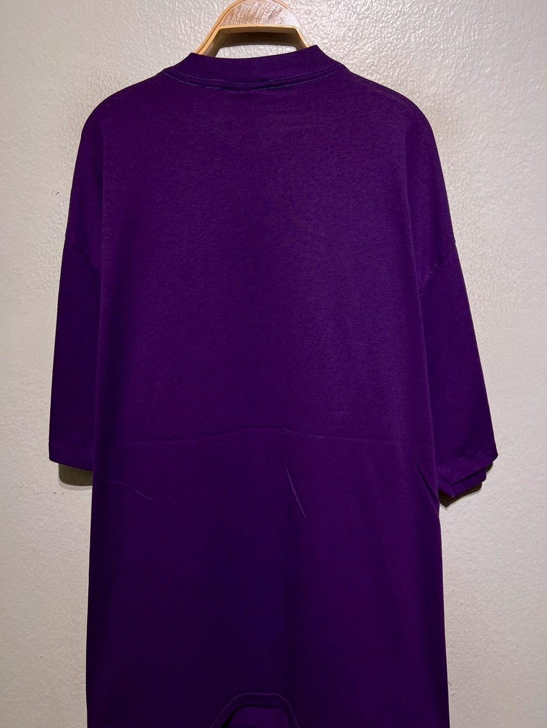 Puede incluir: Mono morado liso con cuello alto y mangas cortas, colgado de una percha de madera. La prenda se exhibe contra una pared neutra. El mono parece estar hecho de un material suave y c&oacute;modo.