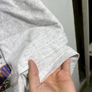 May include: Close-up of a light gray t-shirt sleeve. The sleeve is folded, revealing the hem and a portion of the shirt's graphic design. The fabric has a heathered texture.