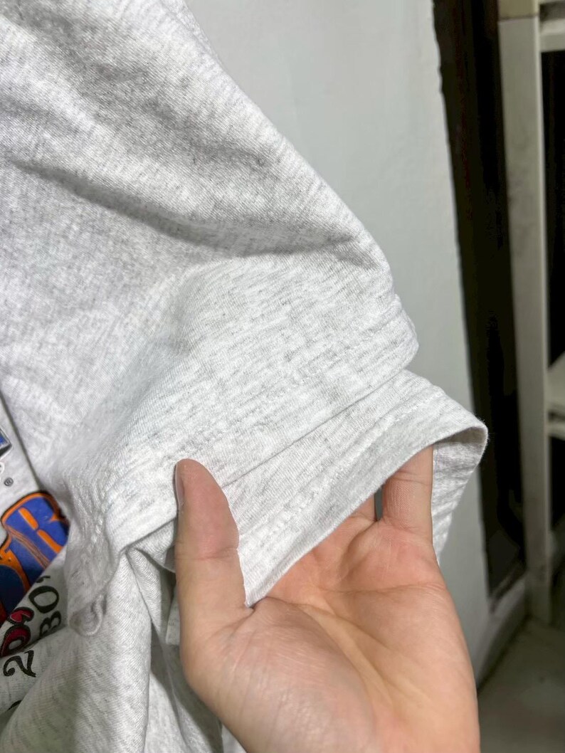 May include: Close-up of a light gray t-shirt sleeve. The sleeve is folded, revealing the hem and a portion of the shirt's graphic design. The fabric has a heathered texture.