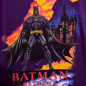 Puede incluir: Camiseta morada con Batman de "Batman Returns". El gr&aacute;fico muestra a Batman con un traje negro con cintur&oacute;n amarillo y el s&iacute;mbolo del murci&eacute;lago, frente a un horizonte urbano y nubes coloridas. Las palabras "BATMAN RETURNS" est&aacute;n impresas en rojo.
