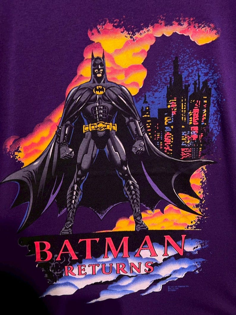 Puede incluir: Camiseta morada con Batman de "Batman Returns". El gr&aacute;fico muestra a Batman con un traje negro con cintur&oacute;n amarillo y el s&iacute;mbolo del murci&eacute;lago, frente a un horizonte urbano y nubes coloridas. Las palabras "BATMAN RETURNS" est&aacute;n impresas en rojo.