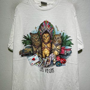 May include: White t-shirt featuring a graphic of three cats, one wearing a feathered headdress, with playing cards, dice, and classic cars. The text "LAS VEGAS" is printed below the cats.
