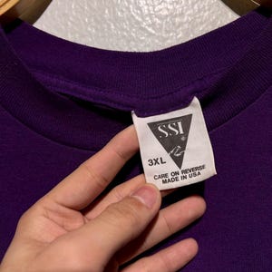 Puede incluir: Primer plano de una camiseta morada oscura con una etiqueta. La etiqueta dice "SSI" con un logotipo, "3XL" y "CARE ON REVERSE MADE IN USA". La camiseta est&aacute; colgada en una percha de madera.