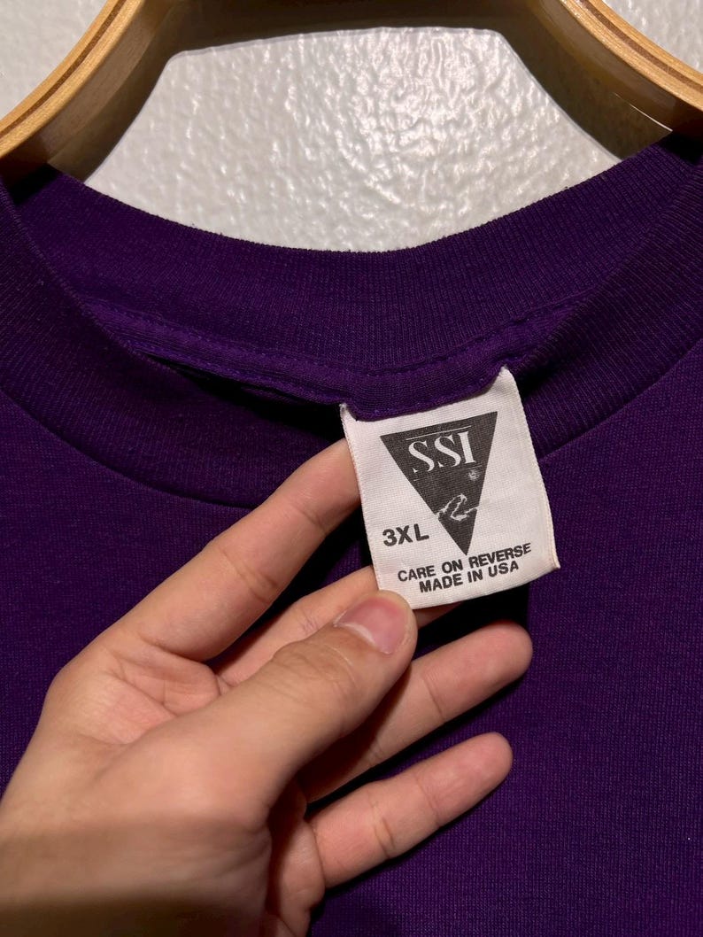 Puede incluir: Primer plano de una camiseta morada oscura con una etiqueta. La etiqueta dice "SSI" con un logotipo, "3XL" y "CARE ON REVERSE MADE IN USA". La camiseta est&aacute; colgada en una percha de madera.
