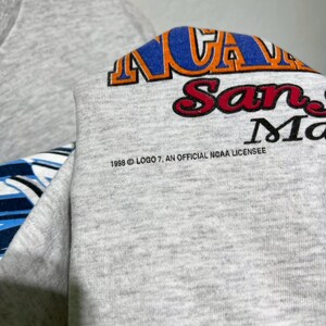May include: A gray sweatshirt with the text "NCAA San Marcos" in blue, orange, and red. The text "1998 Logo 7, An Official NCAA Licensee" is printed below the main text.