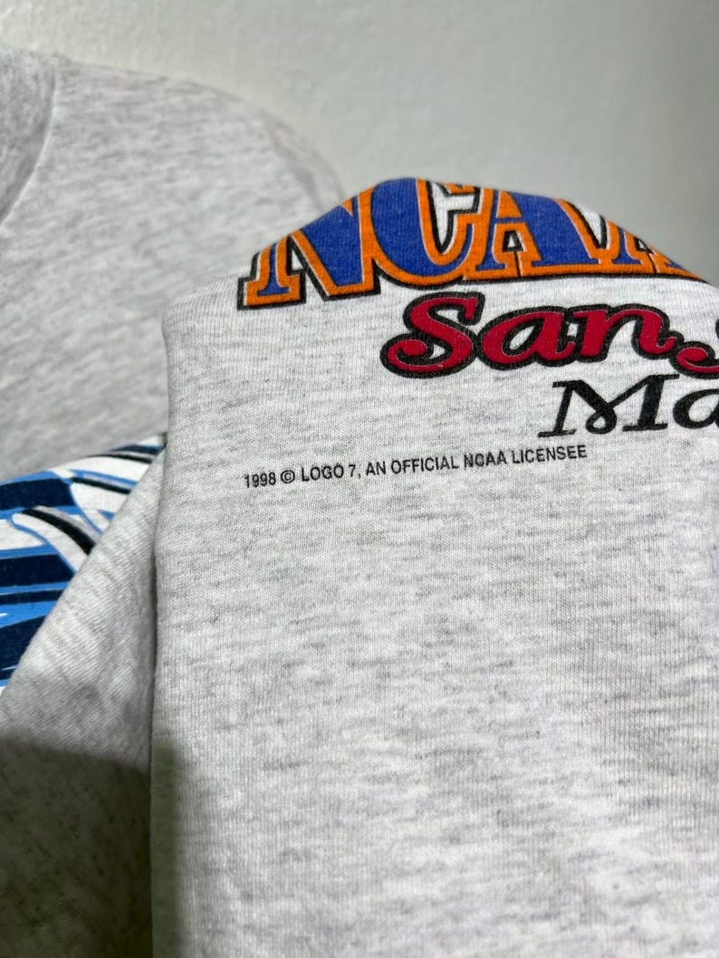 May include: A gray sweatshirt with the text "NCAA San Marcos" in blue, orange, and red. The text "1998 Logo 7, An Official NCAA Licensee" is printed below the main text.