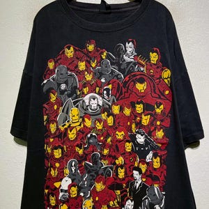 May include: Black t-shirt featuring a collage of Iron Man illustrations in red, yellow, gray, and white. The words "IRON MAN" are printed in white at the bottom. The shirt is hanging on a wooden hanger.