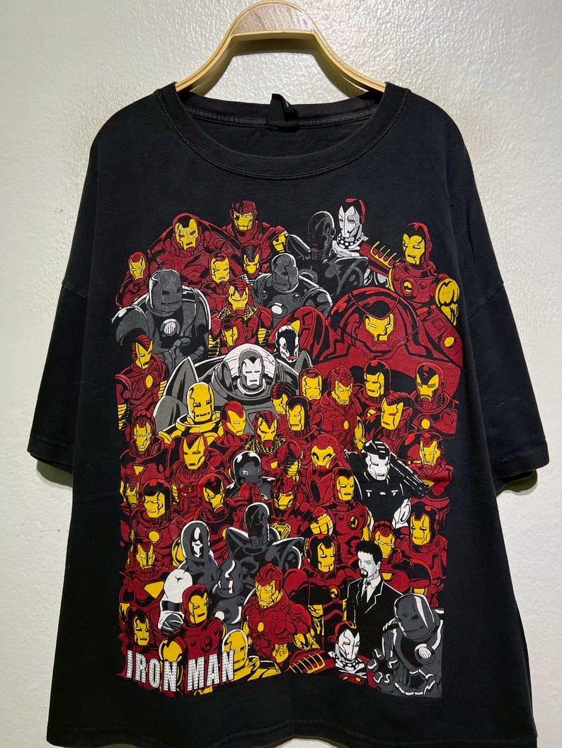 May include: Black t-shirt featuring a collage of Iron Man illustrations in red, yellow, gray, and white. The words "IRON MAN" are printed in white at the bottom. The shirt is hanging on a wooden hanger.