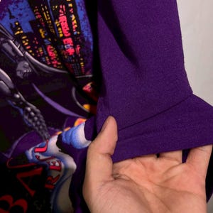 Puede incluir: Camiseta morada con un gr&aacute;fico de Batman. El dise&ntilde;o incluye un paisaje urbano, el logotipo de Batman y la palabra "BATMAN". Se aprecia la textura de la manga, sostenida por una mano.