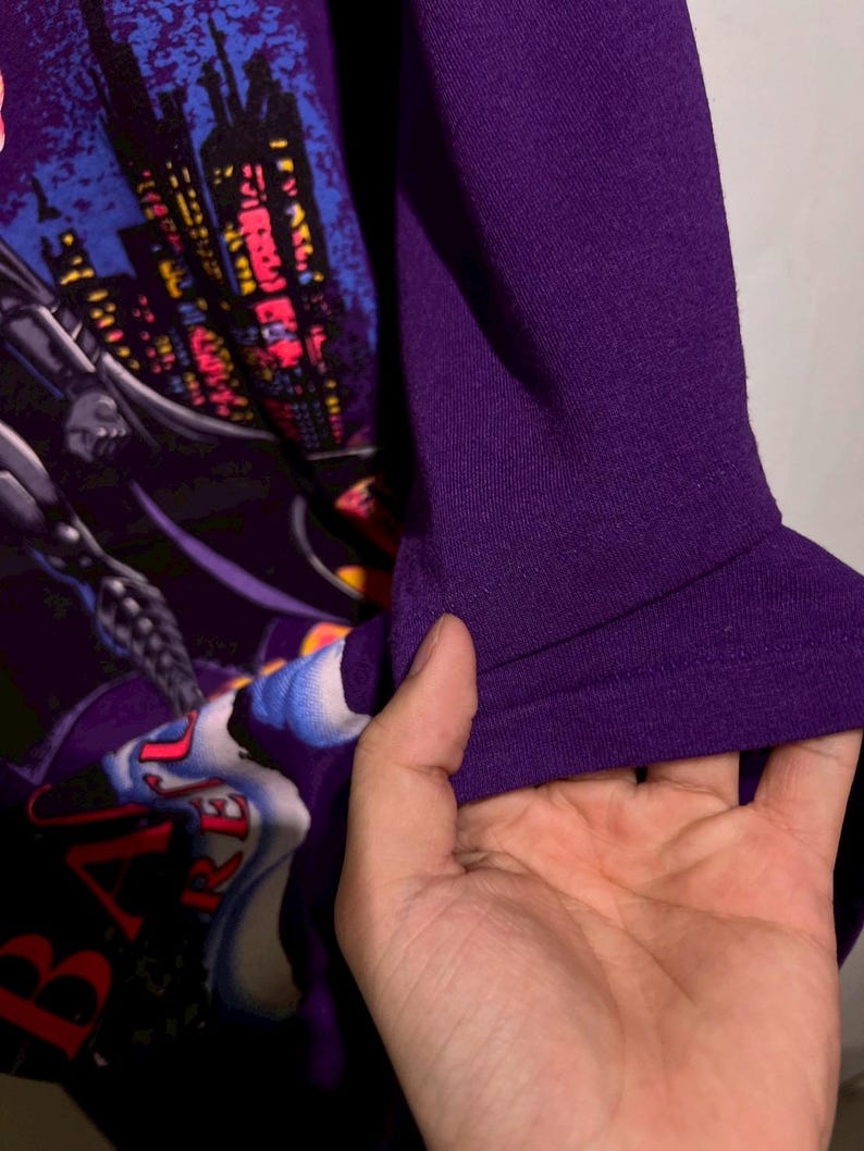 Puede incluir: Camiseta morada con un gr&aacute;fico de Batman. El dise&ntilde;o incluye un paisaje urbano, el logotipo de Batman y la palabra "BATMAN". Se aprecia la textura de la manga, sostenida por una mano.