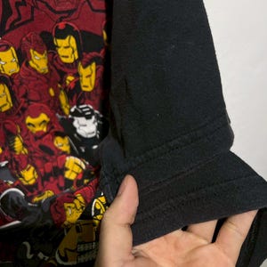 May include: Black and red graphic t-shirt featuring multiple Iron Man helmet designs. The shirt has black sleeves and a crew neck. The graphic is a collage of Iron Man's helmet in various poses and colors.