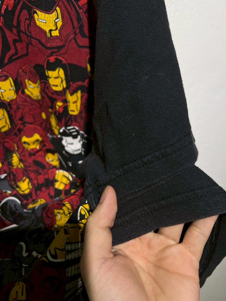 May include: Black and red graphic t-shirt featuring multiple Iron Man helmet designs. The shirt has black sleeves and a crew neck. The graphic is a collage of Iron Man's helmet in various poses and colors.