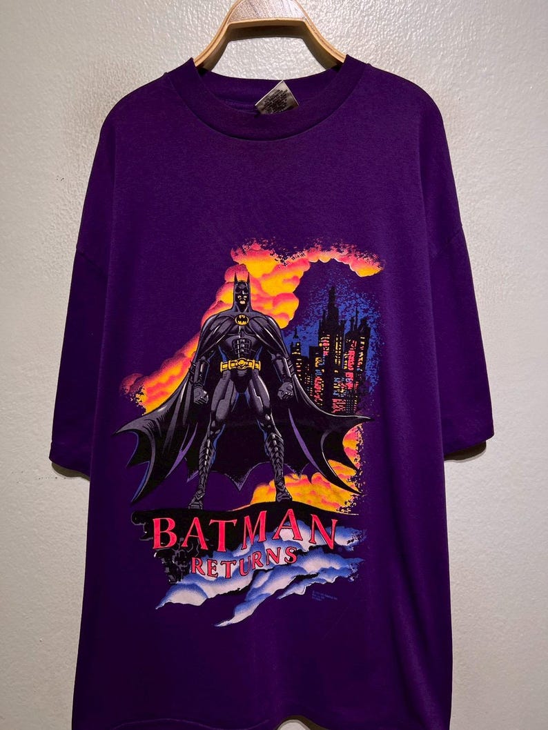 Puede incluir: Camiseta morada de Batman Returns con un gr&aacute;fico de Batman frente a un horizonte urbano con un fondo de puesta de sol. El texto "BATMAN RETURNS" est&aacute; en rosa y blanco.
