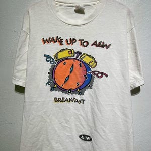 Könnte beinhalten: Weißes T-Shirt mit einer bunten Grafik einer Weckuhr und dem Text "WAKE UP TO A&W BREAKFAST". Das Zifferblatt der Uhr ist orange und blau, mit gelben Alarmglocken. Das A&W-Logo befindet sich unten.