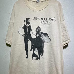 May include: A cream-coloured t-shirt displaying a black and white graphic of two people and the text "Fleetwood Mac Rumours." The graphic shows a man and a woman in a vintage style. The shirt is hanging on a wooden hanger.