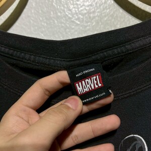 May include: Black t-shirt with a Marvel tag held by a hand. The tag reads "MAD ENGINE" and "MARVEL" in a red and white box. The website www.marvel.com is also visible. The shirt features a graphic of a character.