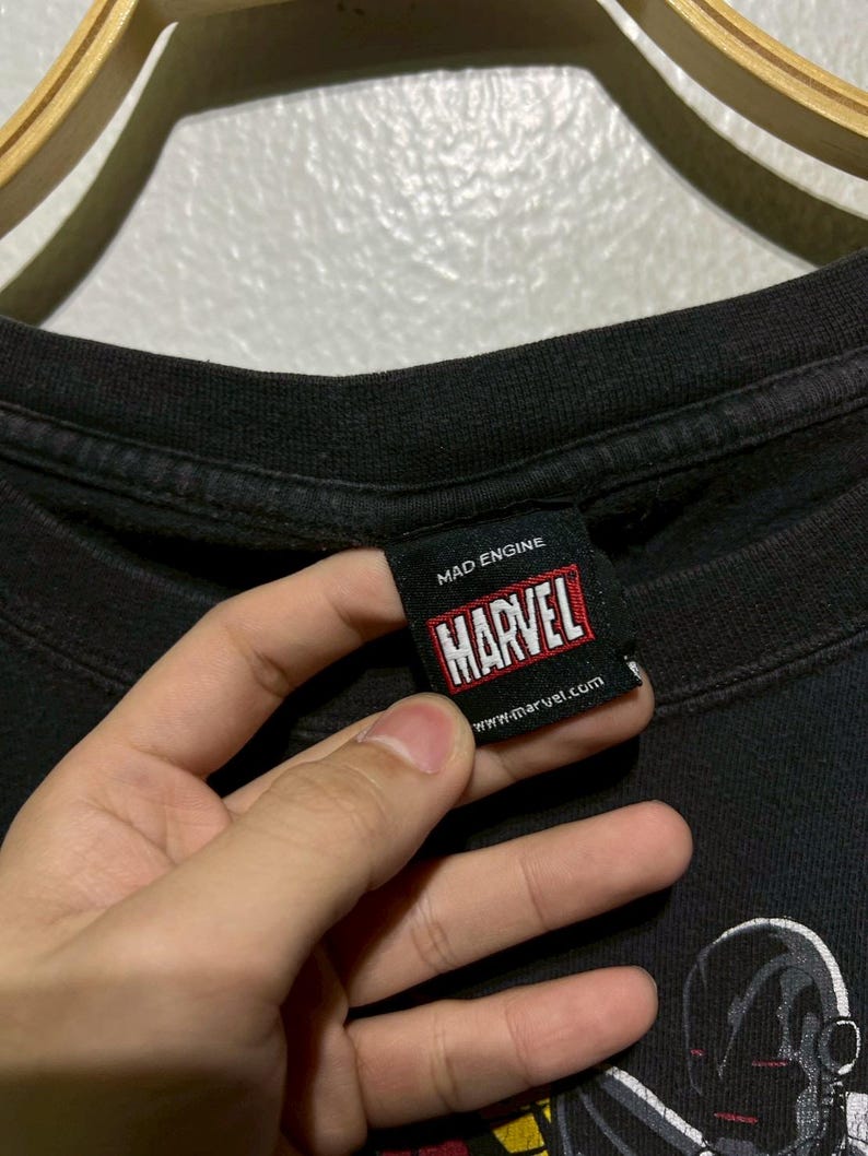 May include: Black t-shirt with a Marvel tag held by a hand. The tag reads "MAD ENGINE" and "MARVEL" in a red and white box. The website www.marvel.com is also visible. The shirt features a graphic of a character.