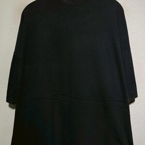 May include: A solid black t-shirt with short sleeves. The shirt is hanging on a wooden hanger against a light gray wall. The fabric appears to be a soft material.