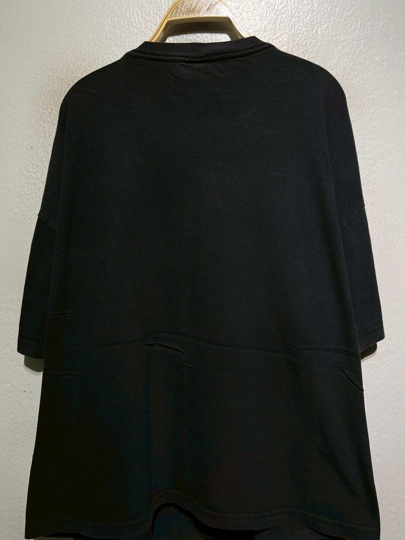 May include: A solid black t-shirt with short sleeves. The shirt is hanging on a wooden hanger against a light gray wall. The fabric appears to be a soft material.
