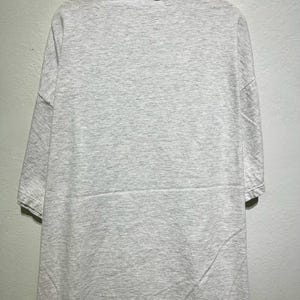 May include: A light gray, short-sleeved t-shirt hanging on a wooden hanger. The shirt has a mock neck and a simple, minimalist design. The fabric appears to be a soft, comfortable material.
